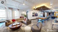 Located at 1000 W Main St in Durham, NC, this single-floor communal space blends industrial-chic design with warm, inviting finishes across a generously sized layout. The living room serves as the centerpiece, featuring exposed ductwork and piping along the ceiling, reclaimed wood beam accents, and an open-plan arrangement that encourages social interaction. Comfortable sectional seating, a large area rug, and bold artwork on white walls create a vibrant yet relaxed atmosphere. A pool table and wall-mounted TV add to the entertainment appeal, while a built-in oversized Scrabble board and oversized Connect Four game provide additional recreational touches. Large windows dressed with sheer curtains flood the space with natural light and offer views of the surrounding Durham streetscape. The kitchen area is outfitted with stainless steel appliances, sleek gray cabinetry, and a warm wood-paneled wall adorned with angular open shelving, perfect for displaying décor. The hallway connects seamlessly to the main areas, finished with wood-look tile flooring. An adjacent outdoor terrace, accessible via full-height glass garage-style doors, features bar-height seating and an outdoor TV, delivering panoramic city views under an open sky.