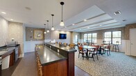 Located at 3595 Owasso St in Shoreview, MN, this communal dining and kitchen space offers a thoughtfully designed single-floor layout that balances functionality with refined aesthetics. The dining area is generously sized, accommodating multiple dining tables and chairs arranged throughout a patterned carpet floor. A coffered ceiling with recessed linear lighting adds architectural character and a sense of depth overhead, while wainscoting and elegant damask-patterned wallcoverings lend a warm, upscale ambiance to the walls. Several windows along the perimeter allow natural light to fill the room and provide views of the surrounding outdoor landscape, complementing the well-lit interior. A wall-mounted flat-screen television serves as a central focal point in the dining area, and high-top seating along the window wall offers an additional casual dining option. The adjoining kitchen is equipped with stainless steel appliances, rich warm-toned cabinetry, and dark granite countertops. A peninsula-style breakfast bar with pendant lighting above and bar stool seating along one side creates a natural transition between the kitchen and dining areas. Hardwood-style flooring grounds the kitchen zone, while glass-paneled doors provide access to adjacent spaces. Together, these areas form a cohesive, welcoming environment well-suited for communal dining and gathering.