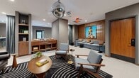 Located at 608 Second Ave S in downtown Minneapolis, this thoughtfully designed amenity space spans a generous single floor and offers a diverse range of social and recreational zones. The main lounge area features large-format terrazzo-style tile flooring, recessed lighting, and a sophisticated neutral color palette accented by warm wood tones. Flexible seating arrangements include plush leather sofas, swivel armchairs, and gold-accented ottomans, creating inviting areas for both casual gatherings and relaxed lounging. A standout entertainment zone is anchored by a built-in wood media wall with integrated shelving and a large display screen. An adjacent game room houses a mid-century modern pool table beneath pendant lighting, accompanied by retro-styled leather chairs and bold framed artwork. A dedicated music and social lounge features a striking disco ball centerpiece, a vinyl record console, mid-century seating, and a vivid large-scale painting set against a wood-paneled accent wall. The bar and dining area includes a stylish curved bar with backlit shelving, bar seating, and a communal dining table suited for group events. Velvet curtain dividers provide flexible spatial separation throughout. Well-curated artwork lines the hallways connecting each zone, reinforcing the space's cohesive, design-forward character.