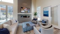 This thoughtfully designed single-floor clubhouse offers a sophisticated and welcoming environment for community members to gather, relax, and entertain. The open-plan layout centers around a spacious lounge area anchored by a dramatic floor-to-ceiling tiled fireplace wall, complete with a wall-mounted television and comfortable contemporary seating arranged to encourage conversation. Soaring ceilings throughout lend an expansive, airy feel, while large windows invite abundant natural light and views of the surrounding community.

The fully equipped kitchen is a standout feature, showcasing dark cabinetry with sleek hardware, a striking white quartz island with bar stool seating, stainless steel appliances including a professional-grade range and refrigerator, a subway tile backsplash, and open floating shelves for both storage and display. A dedicated dining area nearby provides additional seating for shared meals or events.

The clubhouse also includes a separate lounge nook with upholstered armchairs and blue accent pillows, positioned adjacent to glass-paneled private rooms ideal for meetings or smaller gatherings. Light wood-toned flooring flows seamlessly throughout, tying each zone together with a cohesive, modern aesthetic. Curated artwork in cool blue tones adds a refined finishing touch to the overall ambiance.