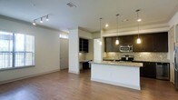 This single-story space showcases an open-concept layout that seamlessly connects the living area and kitchen, creating a cohesive and functional environment. The hardwood flooring runs throughout, adding warmth and continuity to the space.

The kitchen is a standout feature, equipped with dark espresso cabinetry that contrasts elegantly with the light granite countertops. A peninsula with a breakfast bar provides additional seating and workspace, while a stone tile backsplash adds texture and visual interest. Stainless steel appliances, including a range and microwave, are neatly integrated into the design. Pendant lighting hangs above the peninsula, providing focused task lighting that complements the recessed ceiling lights above.

Adjacent to the kitchen is a built-in desk area with open shelving, offering a practical workspace or study nook. The entryway features a transom window above the door, allowing natural light to filter into the space. Track lighting in the living area provides flexible illumination, while crown molding details add a refined architectural touch. The overall ambiance is modern and well-appointed, balancing practicality with contemporary style.