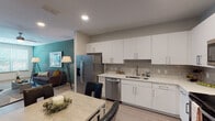 This well-designed single-floor residence offers a thoughtful layout that balances modern aesthetics with everyday functionality. The open-concept kitchen is a standout feature, outfitted with crisp white cabinetry extending to the ceiling, light quartz countertops, a tile backsplash, and a full suite of stainless steel appliances including a French door refrigerator, dishwasher, microwave, and range. Light wood-style flooring flows seamlessly from the kitchen through the adjoining dining area, which comfortably accommodates a full dining table and chairs. The living room is spacious and inviting, anchored by a bold teal accent wall that adds a pop of color against the neutral palette. A ceiling fan, large windows with blinds, and light hardwood floors give the space an airy, comfortable feel. A stylish display cabinet and curated wall art add character to the entryway transition area. The bedrooms are carpeted and well-proportioned, each featuring windows that bring in natural light, ceiling fans, and ample room for furnishings. The bathroom is clean and modern, with large-format gray tile surrounding the soaking tub and shower combination, complemented by marble-look tile flooring and brushed nickel fixtures throughout.