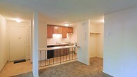 This single-floor space is designed with an open, functional layout that connects the kitchen and living areas seamlessly. The kitchen is outfitted with warm medium-toned wood cabinetry mounted above a clean white countertop backsplash, providing ample upper storage. Essential appliances, including a range and dishwasher, are neatly integrated into the cabinetry. A peninsula-style breakfast bar extends from the kitchen, framed by a decorative metal railing that visually separates the kitchen from the adjoining living space without fully enclosing it.

The living area features wall-to-wall carpeting in a neutral tone, contributing to a comfortable, understated ambiance. White columns and partition walls define the space while maintaining an open feel throughout. A notable feature visible from the main living area is a dedicated walk-in closet, complete with a full-width shelf and hanging rod, offering practical storage solutions. The ceilings feature a subtle texture, and flush-mount lighting provides consistent illumination throughout. The overall palette of warm neutrals and wood tones creates a cohesive, welcoming atmosphere throughout the space.