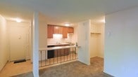 This single-floor space is designed with an open, functional layout that connects the kitchen and living areas seamlessly. The kitchen is outfitted with warm medium-toned wood cabinetry mounted above a clean white countertop backsplash, providing ample upper storage. Essential appliances, including a range and dishwasher, are neatly integrated into the cabinetry. A peninsula-style breakfast bar extends from the kitchen, framed by a decorative metal railing that visually separates the kitchen from the adjoining living space without fully enclosing it.

The living area features wall-to-wall carpeting in a neutral tone, contributing to a comfortable, understated ambiance. White columns and partition walls define the space while maintaining an open feel throughout. A notable feature visible from the main living area is a dedicated walk-in closet, complete with a full-width shelf and hanging rod, offering practical storage solutions. The ceilings feature a subtle texture, and flush-mount lighting provides consistent illumination throughout. The overall palette of warm neutrals and wood tones creates a cohesive, welcoming atmosphere throughout the space.