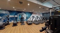 Located at 1714 N Las Palmas Ave in the heart of Hollywood, this contemporary fitness center offers a well-appointed, single-floor workout facility designed to inspire and perform. The space is defined by a bold, athletic aesthetic, anchored by a striking full-wall mural depicting sprinter Usain Bolt alongside the motivational quote, "I know what I can do, so I never doubt myself." Deep navy blue walls contrast with warm wood-look flooring throughout the cardio zone, which is lined with a comprehensive array of treadmills, stair climbers, ellipticals, and stationary and recumbent bikes — all equipped with digital screens. A dedicated strength and free weights area features a full rack of dumbbells, wall-mounted mirrors, and vertical training stations, set on durable carpet tile flooring. The functional training zone is a standout, boasting a tall double-height ceiling that opens above, flooded with natural light from elevated windows. This area includes a Training Zone rig with TRX suspension trainers, kettlebells, medicine balls, BOSU trainers, and sled equipment. A decorative rope installation and a sculptural pendant light add an artistic dimension to the open training floor. Recessed lighting throughout ensures a bright, focused environment across all workout areas.
