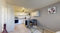 This efficiently designed 2-bedroom, single-floor unit makes smart use of its compact footprint, offering a functional layout that incorporates a combined kitchen and living area along with two bedrooms and a utility storage room. Light beige ceramic tile flooring runs consistently throughout the space, creating a cohesive and easy-to-maintain interior.

The kitchen is well-appointed with white cabinetry, a double-basin sink, granite-style countertops, a full-size refrigerator, and a range. A ceiling fan overhead provides airflow and light, while a dining table nestled nearby offers a convenient eat-in area. The adjacent living room features a wall-mounted TV, a front entry door, and a window with vertical blinds that allows natural light in.

The primary bedroom accommodates a full-size bed and includes a wall-mounted air conditioning unit, a window with curtains, and access to a dedicated walk-in storage room equipped with built-in shelving and a water heater. The second bedroom mirrors the primary in style, with tile flooring and a textured ceiling.

A separate utility room with open shelving and a water heater provides additional storage. Textured ceilings are present throughout, and wall-unit air conditioners ensure climate control in key rooms.