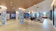 This generously sized amenity space presents a thoughtfully designed, contemporary environment that seamlessly blends social, work, and lounge functions across one expansive floor. Light wood-look flooring extends throughout the open-plan main area, complemented by a warm slatted wood ceiling detail in the entry zone that adds texture and architectural interest.

The main lounge is anchored by multiple curated seating vignettes, featuring modern sofas, lounge chairs, and area rugs that define distinct zones within the open layout. A large-format wall-mounted display serves as a focal point, while original artwork and striking sculptural installations—including a dramatic line-art wrapped column and a tall wooden figurative sculpture—lend a gallery-like quality to the space.

A dedicated kitchenette area offers a long communal table with ample seating beneath bold pendant lighting, stainless steel appliances, a coffee station, and a wall-mounted TV framed by geometric tile. Adjacent lounge nooks provide quieter settings with abundant natural light flowing in through large windows that frame lush outdoor views.

A private conference room with floor-to-ceiling windows, a round table, and a sculptural pendant light offers an intimate setting for focused meetings. Throughout, the design balances openness with defined spaces, creating a dynamic and welcoming environment.
