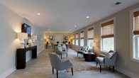 Located at 1751 Dogwood Drive in Charleston, South Carolina, this single-story space offers a thoughtfully designed open-concept floor plan that seamlessly connects the living and dining areas. The living area is generously proportioned, featuring recessed lighting, neutral-toned walls, and light tile flooring that runs throughout, creating a cohesive and airy feel. Comfortable upholstered seating arranged around a round wood accent table anchors the living space, complemented by a woven area rug that adds warmth and texture. A row of large windows dressed with natural bamboo Roman shades lines one wall, allowing abundant natural light to flood the interior while maintaining a sense of privacy. Framed artwork decorates the walls, adding character and a curated aesthetic. The dining area flows naturally from the living space and features a clean-lined table with modern white chairs, flanked by additional framed pieces and greenery accents. Recessed ceiling lights provide consistent, even illumination throughout. The overall ambiance is calm, neutral, and welcoming, with a coastal-inspired design sensibility that reflects the Charleston locale — making it a versatile and functional space suited for a variety of uses.