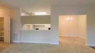 This single-floor space is thoughtfully designed with an open-concept layout that promotes a seamless flow between living areas. The kitchen is positioned behind a half-wall pass-through, offering a practical and social design that keeps the cook connected to the surrounding spaces. White cabinetry lines the kitchen walls, complemented by white appliances and overhead fluorescent lighting that ensures a bright, functional workspace. The countertop pass-through also provides an opportunity for casual seating or serving.

Adjacent to the kitchen, a spacious room is anchored by a classic chandelier with candelabra-style lights, adding a touch of elegance to the otherwise understated interior. This area is finished with neutral wall-to-wall carpeting in a warm beige tone, which runs consistently throughout the visible spaces, contributing to a cohesive and comfortable ambiance. The walls are painted in soft, neutral tones, creating a calm and inviting atmosphere. Clean white baseboards and trim provide crisp definition throughout. Overall, the space offers a clean, move-in ready environment with a traditional yet versatile aesthetic suited to a variety of furnishing styles.