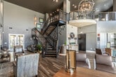 This impressive community clubhouse or leasing center showcases a bold, contemporary design anchored by soaring double-height ceilings that flood the space with natural light through expansive clerestory windows. Rich wood-look flooring runs throughout, providing a warm contrast to the cool gray walls and dark architectural accents. At the heart of the space, a dramatic black steel spiral staircase serves as a sculptural centerpiece, rising to an open mezzanine level above with cable-rail balustrades offering views down to the main floor below. The entry area features floor-to-ceiling glass doors and a dedicated reception desk, creating a welcoming and professional first impression. A branded accent wall displays a prominent oval logo sign flanked by matching lamps atop a white distressed console table, reinforcing a polished, cohesive identity. Multiple seating zones are thoughtfully arranged throughout, including leather lounge chairs and a sofa grouping, offering comfortable spaces for relaxation or conversation. Recessed nooks with dark shiplap paneling house wall-mounted displays and decorative shelving, adding depth and visual interest. Pendant lighting and wall sconces complement the recessed overhead lighting, contributing to a layered, inviting ambiance. Large indoor plants and curated artwork add organic warmth to this refined, upscale environment.