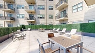 Located at 383-389 Main St, this outdoor terrace is a well-appointed amenity space designed for relaxation and social gathering. Set within a modern multi-story residential building, the terrace features a spacious open-air layout finished with large-format light grey porcelain pavers that lend the space a clean, contemporary aesthetic.

The terrace is thoughtfully furnished with multiple white chaise lounges arranged in clusters, ideal for sunbathing and leisurely outdoor enjoyment. Complementing the lounge seating are several dining and café-style table sets with mixed wood and metal chairs, providing dedicated areas for outdoor dining or casual conversation.

A defining architectural feature of the terrace is the continuous lush green hedge wall that lines the perimeter, creating a sense of privacy while softening the look of the surrounding building facade. Large glass sliding doors and entry points connect the terrace directly to the building's interior, ensuring seamless indoor-outdoor flow.

The surrounding multi-story building rises above on multiple sides, with residential balconies overlooking the terrace, while one open edge of the terrace reveals expansive city skyline views under an open sky. The overall ambiance is modern, bright, and inviting — a well-designed outdoor retreat suited for both quiet relaxation and social gatherings.