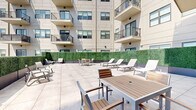 Located at 383-389 Main St, this outdoor terrace is a well-appointed amenity space designed for relaxation and social gathering. Set within a modern multi-story residential building, the terrace features a spacious open-air layout finished with large-format light grey porcelain pavers that lend the space a clean, contemporary aesthetic.

The terrace is thoughtfully furnished with multiple white chaise lounges arranged in clusters, ideal for sunbathing and leisurely outdoor enjoyment. Complementing the lounge seating are several dining and café-style table sets with mixed wood and metal chairs, providing dedicated areas for outdoor dining or casual conversation.

A defining architectural feature of the terrace is the continuous lush green hedge wall that lines the perimeter, creating a sense of privacy while softening the look of the surrounding building facade. Large glass sliding doors and entry points connect the terrace directly to the building's interior, ensuring seamless indoor-outdoor flow.

The surrounding multi-story building rises above on multiple sides, with residential balconies overlooking the terrace, while one open edge of the terrace reveals expansive city skyline views under an open sky. The overall ambiance is modern, bright, and inviting — a well-designed outdoor retreat suited for both quiet relaxation and social gatherings.