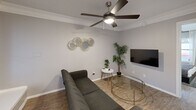Located at 5200 N Lamar Blvd in Austin, TX, this efficient one-bedroom, one-bathroom unit offers a thoughtfully designed layout across a single floor. Light gray walls and wood-look flooring run throughout, creating a cohesive and clean aesthetic.

The living room features crown molding, a ceiling fan with integrated lighting, and a wall-mounted flat-screen TV. The space is open enough to comfortably accommodate a sofa and coffee table, making it a functional area for relaxing or entertaining.

The kitchen is well-appointed with white shaker-style cabinetry, white countertops, a gray tile backsplash, a double-basin stainless steel sink, and black appliances including a range and dishwasher. A pantry closet adds practical storage.

The bedroom is a standout space, highlighted by a bold textured accent wall that adds depth and character. A ceiling fan, a dedicated workspace with a desk and chair, a natural light window with curtains, and two closets round out the room's functionality.

The bathroom includes a combined tub and shower with white tile surround and brushed nickel fixtures, maintaining the clean, neutral palette found throughout the unit.

With two closets providing ample storage, this unit maximizes every square foot, making it a practical and stylish home in the heart of Austin.
