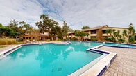 This outdoor communal space centers around a generously sized swimming pool with vivid turquoise water, finished with a distinctive cobalt blue tile border. The pool's irregular shape includes several built-in step platforms and entry points, with stainless steel handrails providing convenient access. Depth markers are clearly visible, and a no-diving policy is posted, reflecting a well-maintained and safety-conscious environment.

Surrounding the pool, a broad paver-stone deck provides ample space for lounge chairs and relaxation. A standout feature is the thatched-roof gazebo situated at one end of the pool area, housing a shaded bench seating area ideal for gathering. Mature oak trees and lush tropical plantings, including palms, crotons, yuccas, and neatly trimmed hedgerows, create a verdant, resort-like atmosphere throughout the grounds.

The complex buildings feature a combination of brick and light-toned siding with pitched rooftops, balconies, and large windows that overlook the pool courtyard. Gravel-mulched garden beds with tropical flora line the building perimeters, enhancing the overall landscaped aesthetic. The cohesive design of the outdoor area evokes a relaxed, Florida-style resort ambiance.