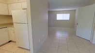 This single-story space presents a straightforward and functional layout designed for comfortable everyday living. The interior is finished with large-format light-toned ceramic tile flooring that runs throughout, creating a cohesive and easy-to-maintain surface underfoot. The open living area is bright and airy, benefiting from natural light admitted through a window that faces the main living space, giving the room a welcoming atmosphere. Neutral beige walls provide a versatile backdrop throughout the space, lending it a clean and understated aesthetic. The kitchen is positioned adjacent to the living area and is equipped with white cabinetry and a full-size refrigerator, offering practical storage and workspace. A solid entry door with a window provides both access and additional natural light at the far end of the living space. The layout flows openly from the kitchen into the main living zone, making the space feel connected and functional. White baseboards frame the tile floors neatly, adding a finished look to the interior. Overall, the space offers a tidy, utilitarian design that is well-suited for a variety of uses, with its light color palette and tile surfaces contributing to a clean and low-maintenance environment.