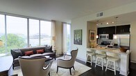 This well-appointed single-floor residence showcases a thoughtfully designed open-concept layout that seamlessly connects the living area with the kitchen. The living space is anchored by large floor-to-ceiling windows that flood the room with natural light and frame expansive views of the surrounding landscape, creating a bright and airy ambiance throughout. Elegant sheer curtains frame the windows, softening the light while maintaining the connection to the outdoors.

The living area features rich dark hardwood flooring that contrasts beautifully with the neutral-toned walls and ceiling. Comfortable seating, including a plush sofa and accent chairs, is arranged around a patterned area rug, creating an inviting gathering space. Artistic wall décor adds a contemporary touch to the room.

The kitchen is open to the living area and is equipped with dark cabinetry, stainless steel appliances, and a stone-topped breakfast bar with wicker bar stools. Pendant lighting above the bar adds warmth and definition to the space. The overall design strikes a balance between modern sophistication and livable comfort, with a cohesive color palette of dark wood tones, soft neutrals, and warm accent hues that ties the entire space together.