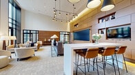 Located at 300 Alexander Ct in Philadelphia, PA, this single-floor amenity space offers a generously sized, thoughtfully designed environment spanning over 2,500 square feet. The layout is organized into distinct yet interconnected zones, each contributing to an upscale, community-oriented atmosphere.

The lounge area commands attention with soaring double-height ceilings and dramatic floor-to-ceiling windows framed in dark steel, flooding the interior with natural light and offering views of a landscaped outdoor terrace. Plush seating arrangements — including curved sofas, swivel armchairs in warm neutral tones, and accent tables — create inviting conversational clusters. A striking wall display of decorative gold sunburst mirrors adds an artistic focal point above a secondary seating nook.

A central bar counter with leather-and-metal barstools serves as a social hub, complemented by pendant lighting and a wall-mounted screen framed by warm wood paneling with built-in display shelving. Adjacent to this, a glass-partitioned corridor with track lighting leads to a dedicated fitness area equipped with exercise machines and hardwood flooring.

Throughout, the design palette balances warm wood tones, neutral carpeting, and white walls, unified by carefully curated lighting — from sculptural chandelier clusters to sleek recessed fixtures — to create a refined yet welcoming communal retreat.