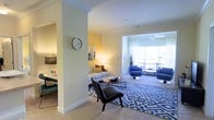 This single-floor residence offers a thoughtfully arranged open layout that connects multiple living areas in a seamless flow. The main living room is generously sized, featuring plush wall-to-wall carpeting and large windows that allow abundant natural light to fill the space, creating a bright and airy atmosphere. A bold blue-patterned area rug anchors the seating arrangement, which includes a light grey sofa, sleek accent chairs, and a round coffee table. Blue upholstered chairs near the window provide an additional relaxed seating nook bathed in natural light, with views to an outdoor area beyond.

Warm yellow accent walls add a cheerful, inviting tone throughout, complemented by soft white crown molding and trim that lend a polished, classic finish. A partial wall with decorative column detailing subtly separates the living room from an adjacent workspace or study area, which features built-in desk space and task lighting. The overall ambiance is modern yet comfortable, with a carefully curated mix of furnishings and warm lighting that makes the space feel both functional and welcoming.