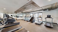 Located at 1000 East Apache Boulevard in Tempe, Arizona, this fitness center offers a comprehensive workout facility spread across two distinct exercise rooms on a single floor. The space features a striking industrial-modern aesthetic, with exposed concrete ceilings complemented by sleek linear pendant lighting that runs the length of the main cardio room, creating a dynamic and energizing atmosphere.

The primary cardio room is generously proportioned, housing an extensive lineup of Matrix treadmills, stair climbers, and elliptical machines arranged in parallel rows. Full-length mirrors along the walls visually expand the space and allow users to monitor their form, while large floor-to-ceiling windows flood the room with natural light and provide views of the surrounding Tempe urban landscape. Neutral-toned flooring completes the clean, functional aesthetic.

The second exercise room offers a distinct environment tailored for functional and cross-training workouts. It features warm hardwood-style flooring, a full mirror wall, and an array of equipment including spin bikes, medicine balls, kettlebells, dumbbells, and resistance training accessories. Glass partition walls with black steel framing connect the two rooms while maintaining a sense of openness. Together, the two rooms provide a well-rounded fitness facility suited for a wide range of training styles and fitness goals.