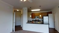This single-floor residence offers a thoughtfully designed open-concept layout that seamlessly connects the kitchen and living areas. The kitchen is a standout feature, showcasing warm honey-toned wood cabinetry that extends to the upper walls, complemented by sleek stainless steel appliances including a refrigerator, range, and built-in microwave. A peninsula with dark granite-style countertops serves as a breakfast bar, providing additional seating and workspace while visually anchoring the space. Pendant lights hang above the bar, adding a touch of warmth alongside under-cabinet and recessed lighting that keeps the area bright and functional. Crown molding details on the kitchen columns add an architectural refinement to the space. The adjacent living area features carpeted flooring and a ceiling fan, creating a comfortable, relaxed atmosphere. A hallway leading to the front entry is visible, featuring tile or wood-look flooring and a classic paneled entry door, offering a welcoming transition into the home. Throughout the space, neutral wall tones and consistent design elements create a cohesive, inviting ambiance suited for everyday living.