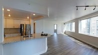 This single-floor residence showcases a thoughtfully designed open-concept layout that seamlessly connects the kitchen and living areas. The kitchen is outfitted with granite countertops featuring subtle veining, a stainless steel refrigerator, and contemporary white cabinetry, all anchored by a generous peninsula that serves as both a prep space and casual gathering point. A modern sink with a sleek faucet is integrated into the peninsula, adding both function and visual interest.

The flooring throughout is a warm-toned hardwood-style material that runs continuously from the kitchen into the expansive living area, reinforcing the cohesive, open feel of the space. White walls and recessed lighting in the kitchen zone contribute to a clean, bright aesthetic. The living area benefits from multiple windows that invite substantial natural light, creating an airy atmosphere. A hallway visible in the background suggests additional rooms beyond the main living space. The overall design balances modern finishes with a neutral palette, resulting in a versatile and inviting environment.