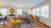 Located at 597 Millville Ave in Naugatuck, Connecticut, this multi-floor residence spans three levels and offers a welcoming, thoughtfully designed interior. The living room is a standout space, featuring warm yellow walls, hardwood flooring, and an abundance of natural light streaming through multiple windows fitted with blinds. A vibrant striped area rug anchors the seating area, complemented by a gray sofa, bold yellow accent chair, and decorative artwork, creating an upbeat and inviting atmosphere. The living area flows seamlessly into a dedicated dining space, which features a glass-top table with navy chairs beneath a ceiling fan, with easy access to the kitchen. The galley-style kitchen is outfitted with white cabinetry, dark countertops, tile flooring, and full appliances, offering ample storage and workspace. The bedrooms are carpeted and comfortably sized; the primary bedroom features a ceiling fan, generous dresser storage, and tasteful décor, while a secondary bedroom accommodates twin beds and benefits from natural light through a double window. A staircase connects all three floors, offering a practical and well-organized layout throughout. The overall ambiance is bright, clean, and move-in ready.