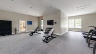 Located in Murfreesboro, Tennessee, this dedicated fitness center offers a comprehensive workout environment designed to accommodate a wide range of exercise routines. The space is finished with durable speckled rubber flooring throughout, providing a safe and resilient surface suitable for both cardio and strength training activities.

The room is organized into distinct training zones. A cardio section houses multiple treadmills positioned along a windowed wall, allowing natural light to fill the space while a wall-mounted flat-screen television provides entertainment during workouts. An additional area features elliptical trainers and a stationary spin bike for low-impact cardiovascular exercise.

The strength training section is well-appointed with a multi-function cable machine, adjustable weight benches, and a tiered dumbbell rack stocked with a full range of free weights. A large floor-to-ceiling mirror spans one wall, visually expanding the space and allowing users to monitor their form. Alongside the mirror, a dedicated functional training zone holds an assortment of medicine balls in various sizes, resistance bands, stability balls, and exercise mats neatly organized on storage racks.

Recessed lighting throughout the ceiling provides consistent, even illumination across the entire floor. Windows on multiple walls invite natural light and views of the surrounding greenery. The clean, neutral color palette and modern finishes give the facility a polished, professional atmosphere.
