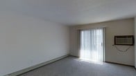 This single-floor space offers a clean, functional layout designed for comfortable everyday living. The main living area is finished with wall-to-wall carpeting in a neutral gray tone, creating a soft and cohesive foundation throughout the room. A textured ceiling adds a classic touch to the interior, while the smooth, white walls provide a bright and open feel.

A standout feature of the living area is the full-width sliding glass door, dressed with vertical blinds that allow for flexible light control. When open, the blinds invite generous amounts of natural light to fill the room, enhancing the overall sense of openness and airiness. The sliding door also provides convenient access to an outdoor area, extending the functional living space beyond the interior.

Along the base of one wall, a baseboard heating unit ensures efficient climate control throughout the colder months, blending seamlessly with the room's understated aesthetic. The neutral color palette of whites, grays, and beiges throughout the space creates a versatile backdrop suitable for a variety of furnishing styles and personal tastes. Overall, this space presents a practical and well-lit environment with key features that support both comfort and functionality.