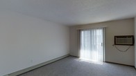 This single-floor space offers a clean, functional layout designed for comfortable everyday living. The main living area is finished with wall-to-wall carpeting in a neutral gray tone, creating a soft and cohesive foundation throughout the room. A textured ceiling adds a classic touch to the interior, while the smooth, white walls provide a bright and open feel.

A standout feature of the living area is the full-width sliding glass door, dressed with vertical blinds that allow for flexible light control. When open, the blinds invite generous amounts of natural light to fill the room, enhancing the overall sense of openness and airiness. The sliding door also provides convenient access to an outdoor area, extending the functional living space beyond the interior.

Along the base of one wall, a baseboard heating unit ensures efficient climate control throughout the colder months, blending seamlessly with the room's understated aesthetic. The neutral color palette of whites, grays, and beiges throughout the space creates a versatile backdrop suitable for a variety of furnishing styles and personal tastes. Overall, this space presents a practical and well-lit environment with key features that support both comfort and functionality.