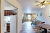 This thoughtfully designed single-floor unit offers a comfortable and functional layout across one bedroom, one bathroom, a living room, and a kitchen. Light wood-look flooring flows consistently throughout the main living areas, creating a cohesive and clean aesthetic.

The kitchen features rich, dark wood cabinetry offering ample storage, white appliances including a coil-top electric range and a refrigerator, and light-toned countertops. An arched pass-through opening connects the kitchen to the dining and living areas, promoting an open, connected feel.

The living and dining area is generously proportioned, illuminated by a ceiling fan with integrated lighting and natural light from a curtained window overlooking a peaceful outdoor setting. The space comfortably accommodates a dining table, seating area, and additional accent chairs.

The bedroom is spacious and well-lit, featuring two closets, a built-in desk area, durable wood-look flooring, and a window with outdoor views that fill the room with natural light.

The bathroom is efficiently designed with a wide vanity, large mirror, tile flooring, and grab bars in the toilet and shower areas, indicating accessibility-conscious construction.

Overall, the unit presents a warm, practical living environment with consistent finishes and thoughtful design throughout.