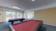 Located at 2370 Lucretia Ave in San Jose, California, this single-floor community recreation facility is designed to provide residents with a dedicated space for leisure and social activity. The space is divided into two main areas that flow openly into one another, creating a versatile and welcoming environment.

The billiards room serves as the centerpiece of the facility, housing two full-sized pool tables with rich red felt surfaces, complemented by a wall-mounted cue rack and seating along the perimeter. Colorful billiard ball-themed artwork adds a playful, thematic touch to the walls. The room features a textured ceiling with recessed fluorescent lighting and is finished with durable carpeted flooring throughout.

Adjacent to the billiards area is a dedicated ping-pong room equipped with a regulation-sized table tennis table. Large sliding glass doors and floor-to-ceiling windows line the walls, flooding the space with abundant natural light and offering unobstructed views of the outdoor deck and mature trees beyond.

A comfortable lounge nook near the sliding doors features upholstered armchairs and a side table, providing a relaxed spot to socialize or unwind. The facility also includes an emergency exit and is well-maintained throughout, making it a functional and enjoyable amenity for the surrounding residential community.