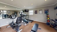 Located at 2842 E 51st St in Tulsa, Oklahoma, this dedicated fitness center offers a well-equipped exercise space designed to support a well-rounded workout routine. The room features neutral gray walls, patterned carpeted flooring, and a textured ceiling with recessed lighting and crown molding that adds a polished finish to the space. Crown molding runs along the perimeter, lending a refined touch throughout.

The room is outfitted with a solid selection of cardio equipment, including treadmills, elliptical trainers, and a stationary bike, primarily from the TRUE Fitness brand. A multi-tier dumbbell rack provides free weight options for strength training, complemented by a flat workout bench. A large wall mirror on one side visually expands the space and supports proper form during exercise. A wall-mounted television adds convenience for entertainment during workouts.

Natural light filters in through a sliding glass door and a window, offering outdoor views of the surrounding landscaped grounds. A series of framed instructional workout posters—covering muscle groups such as chest, core, back, and legs—line the walls, providing helpful exercise guidance. Motivational signage reinforces an encouraging atmosphere. The space is compact yet thoughtfully organized, making efficient use of every square foot for a complete fitness experience.