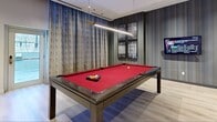 The Games Room and Sport Simulator at Citizen on Jasper is a thoughtfully designed amenity space that combines active recreation with comfortable social lounging. The space is divided into two distinct areas connected by a light-filled corridor finished with light wood-look flooring and clean white walls throughout.

The main games room features a bold design aesthetic, with a striking striped blue accent wall as a dramatic backdrop to a full-sized billiards table with a vibrant red felt surface. A wall-mounted flat-screen TV, stylish pendant lighting, and a lounge area with contemporary white chairs and marble-top tables create a welcoming environment for socializing. Framed game-themed artwork adds a curated, urban character to the space. Large windows dressed with floor-length drapes allow natural light to fill the room, with views of the surrounding cityscape.

The adjacent sport simulator room is dedicated to golf enthusiasts, featuring a professional-grade golf simulator complete with a large projection screen, synthetic turf hitting mat, and a full set of clubs. A generously sized window with elegant draping brings in natural light, complementing the immersive simulator experience. A wall-mounted TV in the connecting hallway adds additional entertainment options. Together, these two areas offer a well-rounded, premium recreational experience for residents.