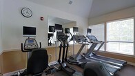 This fitness center is thoughtfully designed to support a well-rounded cardio workout experience. The space is outfitted with a selection of cardio equipment, including treadmills and elliptical trainers, each equipped with individual mounted screens for entertainment or workout tracking during exercise sessions. A large wall mirror spans a significant portion of the room, creating a sense of openness and allowing users to monitor their form while training. Natural light filters in through slatted blinds on the windows, contributing to a bright and energizing atmosphere. The walls feature a neutral warm tone with a two-tone treatment — a lighter upper half and a subtle lower accent — giving the space a clean, welcoming appearance. The flooring is carpeted, providing a degree of comfort underfoot. A wall clock and a fire safety alarm are also visible, reflecting attention to both convenience and safety. The vaulted ceiling adds a sense of airiness to the room, enhancing the overall comfort of the environment. This single-floor fitness space is functionally laid out to maximize equipment accessibility while maintaining an open, uncluttered feel suitable for focused exercise.