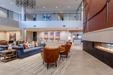 This expansive amenity lounge is defined by its striking architectural presence, anchored by soaring double-height ceilings and a dramatic floor-to-ceiling fireplace surround clad in rich, textured corten-style panels with an abstract linear design. Large-format hexagonal stone tiles flow throughout the main lounge, grounding the space with a clean, neutral palette. A cascading linear chandelier composed of slender metallic rods draws the eye upward, complementing the open upper-level mezzanine railing visible above.

The lounge area is thoughtfully furnished with deep navy sectional sofas, cognac leather armchairs, and mixed-material side tables, creating layered seating arrangements suited for both social gatherings and relaxed conversation. Curated artwork lines the walls throughout, adding vibrant color and visual interest. A glass-walled enclosure adjacent to the main lounge offers a more intimate seating option with outdoor views.

The catering kitchen features a substantial white stone waterfall island, dark cabinetry with stainless steel fixtures, subway tile backsplash, and a slatted wood ceiling detail that adds warmth and texture. It opens directly into a dining area with tufted banquette seating, round tables, and natural light from large windows overlooking greenery, creating an inviting space for private dining and events.