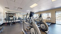 Located at 2100 North 10th Street in El Centro, California, this fitness center offers a comprehensive and thoughtfully arranged exercise facility within a single, open-plan room. The space features a drop-panel ceiling with recessed fluorescent lighting that ensures bright, even illumination throughout, complemented by generous natural light streaming in through multiple large windows that offer views of the surrounding outdoor landscape.

The flooring throughout is durable rubber matting, ideal for both cardio and strength training activities. The cardio section is well-stocked with multiple Matrix elliptical cross-trainers, treadmills, and stationary bikes, providing ample options for aerobic workouts. A dedicated strength training area features a fully loaded power rack with plate-loaded barbells, adjustable weight benches, and a rack of dumbbells spanning a wide range of weights, along with medicine balls for functional training.

Full-length mirrors line one wall of the strength zone, enhancing the sense of space and allowing users to monitor their form. Glass partition walls and double-door entryways add a modern, open feel to the layout. A small refreshment station with a standing table and hydration supplies is conveniently positioned near the entrance. The overall ambiance is clean, functional, and professional, making this facility well-suited for a full-body workout regimen.