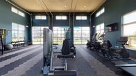 Located at 40000 Argento Way in Kannapolis, NC, this single-floor fitness center is a well-appointed community amenity space designed to support a full range of workout routines. The facility is defined by its dramatic soaring ceilings and bold teal accent walls, creating an energizing and open atmosphere throughout.

The main workout area is anchored by a row of oversized glass garage-style doors that flood the space with natural light while offering unobstructed views of the surrounding green outdoor areas. This section houses a comprehensive selection of Matrix strength-training machines, a flat bench, and a dumbbell rack, catering to resistance and weight training needs.

A dedicated cardio zone lines the perimeter walls, equipped with Matrix treadmills, ellipticals, and upright bikes, each positioned in front of large windows and wall-mounted televisions for an engaging workout experience. Natural light pours in from multiple clerestory windows positioned high along the walls, further enhancing the airy feel of the space.

The functional training area provides stability balls, suspension trainers, medicine balls, and a wall-mounted pull-up rig, offering versatile options for dynamic and bodyweight-focused exercises. Patterned herringbone carpet tiles cover the floors throughout, adding texture and visual interest. The space is completed with recessed lighting and direct outdoor access through glass entry doors.