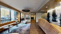 Located at 742 N Aberdeen St in Chicago's vibrant West Town neighborhood, this single-floor lobby space combines industrial-chic architecture with carefully curated interior design. The layout flows openly across wood-plank flooring, anchored by dark cylindrical columns that add structural character throughout.

The main lounge area features multiple distinct seating arrangements, including a large tufted caramel leather sectional sofa positioned beneath floor-to-ceiling windows that frame street-level views of the surrounding neighborhood. Bold botanical-patterned drapery in teal and white accents the glazed exterior walls, while layered area rugs define individual conversation zones. A sculptural woven pendant light fixture and recessed ceiling lighting provide a warm, balanced ambiance.

Textured, marbled wall finishes in muted gray tones run throughout, complemented by dark wood-framed doorways and trim. A striking abstract wall-mounted artwork serves as a focal point along the main corridor. An adjacent dining area features pendant globe lighting and bar-height seating, visible through an open doorway.

A dedicated mailroom and parcel area is equipped with a full bank of individual mailboxes, a mounted display screen, and a stone-topped counter, finished with the same refined material palette found throughout the space.