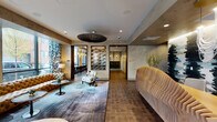 Located at 742 N Aberdeen St in Chicago's vibrant West Town neighborhood, this single-floor lobby space combines industrial-chic architecture with carefully curated interior design. The layout flows openly across wood-plank flooring, anchored by dark cylindrical columns that add structural character throughout.

The main lounge area features multiple distinct seating arrangements, including a large tufted caramel leather sectional sofa positioned beneath floor-to-ceiling windows that frame street-level views of the surrounding neighborhood. Bold botanical-patterned drapery in teal and white accents the glazed exterior walls, while layered area rugs define individual conversation zones. A sculptural woven pendant light fixture and recessed ceiling lighting provide a warm, balanced ambiance.

Textured, marbled wall finishes in muted gray tones run throughout, complemented by dark wood-framed doorways and trim. A striking abstract wall-mounted artwork serves as a focal point along the main corridor. An adjacent dining area features pendant globe lighting and bar-height seating, visible through an open doorway.

A dedicated mailroom and parcel area is equipped with a full bank of individual mailboxes, a mounted display screen, and a stone-topped counter, finished with the same refined material palette found throughout the space.