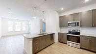 This thoughtfully designed single-story home features three bedrooms, two bathrooms, and a functional open-concept layout. The kitchen serves as the centerpiece of the living area, showcasing a large granite-topped island with an undermount sink, sleek taupe cabinetry with brushed hardware, and a full suite of stainless steel appliances including a refrigerator, range, microwave, and dishwasher. Stylish pendant lights hang above the island, adding a modern touch. The kitchen flows seamlessly into the living room, which features light hardwood-style flooring, recessed lighting, and a wall of windows alongside a glass door that fills the space with natural light and provides outdoor access. The bedrooms are carpeted for comfort and include ceiling fans and windows with blinds. The primary bedroom is a generous size with its own dedicated bathroom, which features granite countertops, a dual-sink vanity with gray cabinetry, a large mirror, and hardwood-style flooring. Additional highlights include three closets, a dedicated laundry area, built-in shelving, and a recessed electrical panel. The clean white walls, contemporary finishes, and cohesive design create a bright, welcoming atmosphere throughout the home.
