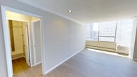 This single-floor space offers a clean, contemporary aesthetic defined by its light hardwood flooring, neutral grey walls, and crisp white trim and baseboards. The main living area is bathed in natural light, courtesy of large floor-to-ceiling windows that frame an urban cityscape, creating an open and airy atmosphere. A baseboard heating unit runs along the window wall, ensuring year-round comfort. Recessed lighting in the ceiling complements the natural light, while elegant crown moulding adds a refined architectural detail that elevates the overall design. The walls are finished in a soft, neutral tone that enhances the sense of brightness and space. An adjacent room is accessible through a white-trimmed doorway, hinting at a well-connected layout. The overall ambiance is minimalist and modern, with attention to clean lines and understated finishes throughout. The combination of abundant natural light, urban views, and polished interior details makes this a sophisticated and functional space suited for a variety of uses.
