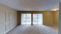 This single-floor residential space offers a clean, functional layout designed for comfortable everyday living. The main living area is a standout feature, characterized by its open and airy feel. Neutral-toned carpeting runs throughout, providing warmth underfoot, while the walls are finished in a soft, earthy tone that lends a welcoming ambiance to the room.

A prominent set of sliding glass doors dominates one wall of the living area, fitted with vertical blinds that allow for adjustable light control. These doors open the room to natural light and provide a direct view of the outdoor surroundings, including a glimpse of trees and the exterior environment beyond. The doors also offer convenient access to an outdoor area, making the transition between indoor and outdoor living seamless.

The ceiling is smooth and finished in crisp white, contributing to the sense of openness throughout the space. Decorative wall molding details add a subtle architectural touch to the otherwise understated interior. The overall design is practical and straightforward, with a neutral palette that offers a versatile canvas for a variety of furnishing styles. This space presents a simple yet functional residential environment suited for single-level living.