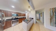 Located at 1517 Cameron Samuel Lane in West Palm Beach, Florida, this single-floor, three-bedroom, two-bathroom home offers a well-designed layout with a comfortable and functional living experience. The open-concept kitchen is a standout feature, showcasing rich walnut-toned shaker cabinetry, dark granite countertops, a central island with a built-in sink, pendant lighting, and a full suite of stainless steel appliances. Light ceramic tile flooring runs throughout the main living areas, lending a clean, cohesive look to the space. The combined dining and living area provides a versatile entertaining space, with the dining room accented by a bold teal feature wall and a large floor mirror that adds depth and brightness. The bedrooms are carpeted for comfort, with the spacious primary bedroom featuring a walk-in closet complete with wire shelving. The bathrooms are well-appointed, with one featuring a tiled tub-shower combo, granite vanity, and warm-toned tile accents. A dedicated laundry room houses a stacked washer and dryer unit for added convenience. The overall ambiance is bright and inviting, reflecting a practical yet stylish Florida residential aesthetic.