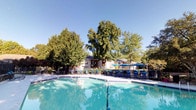 This outdoor amenity space centers around a generously sized swimming pool with clearly marked depth zones, ranging from a shallow entry area to a deeper end, making it suitable for a variety of swimmers. The pool's vivid turquoise water is complemented by a wide concrete pool deck that provides ample room for lounging and socializing. Surrounding the pool, a collection of outdoor chairs and tables offers comfortable seating for residents and guests.

A covered pavilion with blue market umbrellas and additional seating occupies one end of the deck, creating a shaded retreat ideal for relaxing out of direct sunlight. The area is enclosed by a sturdy black wrought-iron fence, ensuring safety and privacy while maintaining open sightlines to the lush, mature trees that frame the space and provide natural shade.

One of the most distinctive architectural features of this pool area is the stacked-stone retaining wall that lines one side of the deck. Constructed from layered natural stone, it adds a refined, rustic character to the space and integrates the pool area seamlessly into the surrounding landscaped grounds. An auxiliary building with a gray entry door is conveniently located poolside, likely serving as a utility or storage facility. Overall, the space offers a welcoming, resort-like atmosphere suited for outdoor recreation and leisure.