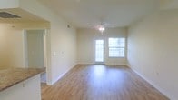 This single-story space in Denton, Texas offers a clean, functional layout with a welcoming open feel. The main living area is highlighted by smooth hardwood-style flooring in a warm honey tone that reflects natural light throughout the room. Neutral beige walls provide a versatile backdrop, complemented by crisp white baseboards that frame the space neatly. A ceiling fan with an integrated light fixture is centrally mounted, offering both ambient lighting and airflow year-round. Natural light enters generously through a glass exterior door fitted with blinds and an adjacent window, creating a bright and airy atmosphere. The layout allows for flexible furniture arrangement within the open living area. A wide doorway or opening visible to one side suggests a connected room or hallway, contributing to the overall flow of the floor plan. The space presents a straightforward, well-maintained interior with consistent finishes throughout, making it a practical and comfortable option in the Denton area.