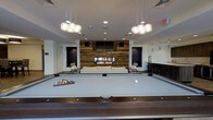 This single-floor amenity lounge is thoughtfully designed to blend modern aesthetics with a welcoming, social atmosphere. At the heart of the space sits a full-sized billiards table, positioned beneath a pair of striking globe pendant light clusters that cast a warm, focused glow over the playing surface. The room's focal point is a reclaimed wood accent wall, which adds rustic warmth and textural contrast against the otherwise clean, white interior. Set within this wall is a wide linear gas fireplace, flanked by a wall-mounted flat-screen television above, and built-in dark niche shelving on either side that provides both display space and visual depth. In front of the fireplace, a plush white sofa and lounge chairs create an inviting seating area, ideal for gathering and conversation. To one side of the room, a wet bar with overhead cabinetry and a counter offers a dedicated space for entertaining. Recessed ceiling lighting complements the pendant fixtures, ensuring the entire space is well-lit and functional. The combination of natural wood elements, contemporary furnishings, and thoughtfully placed lighting gives this lounge a sophisticated yet relaxed character, making it a premier destination for recreation and socializing.