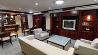 This single-floor space presents a well-appointed living area defined by its classic, professional aesthetic. The room is anchored by an expansive wall unit crafted from rich mahogany-toned wood, featuring open shelving, enclosed cabinetry, and a dedicated media section housing a flat-screen television. Recessed lighting and a central flush-mount ceiling fixture provide warm, even illumination throughout the space, complemented by a ceiling-mounted air conditioning unit for climate control. The seating arrangement centers around a sleek glass-top coffee table with a dark wood frame, surrounded by neutral-toned upholstered armchairs and a generously sized sofa, creating a comfortable yet refined lounging area. The flooring is covered in a textured, patterned carpet that adds depth and warmth underfoot. Toward the rear of the space, a separate dining or work area is visible, furnished with dark wood chairs and a small table, offering a functional secondary zone. The overall ambiance is polished and structured, combining traditional wood detailing with understated contemporary furnishings for a cohesive, hospitality-grade environment.