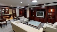 This single-floor space presents a well-appointed living area defined by its classic, professional aesthetic. The room is anchored by an expansive wall unit crafted from rich mahogany-toned wood, featuring open shelving, enclosed cabinetry, and a dedicated media section housing a flat-screen television. Recessed lighting and a central flush-mount ceiling fixture provide warm, even illumination throughout the space, complemented by a ceiling-mounted air conditioning unit for climate control. The seating arrangement centers around a sleek glass-top coffee table with a dark wood frame, surrounded by neutral-toned upholstered armchairs and a generously sized sofa, creating a comfortable yet refined lounging area. The flooring is covered in a textured, patterned carpet that adds depth and warmth underfoot. Toward the rear of the space, a separate dining or work area is visible, furnished with dark wood chairs and a small table, offering a functional secondary zone. The overall ambiance is polished and structured, combining traditional wood detailing with understated contemporary furnishings for a cohesive, hospitality-grade environment.