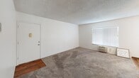 This single-story unit at 553 S Magnolia in El Cajon offers a straightforward, functional layout with a clean, neutral aesthetic throughout. The living area is generously sized, featuring wall-to-wall carpet, white walls, and a textured ceiling. Multiple windows fitted with horizontal blinds allow natural light to fill the space, while a ceiling fan provides additional comfort. A wall-mounted air conditioning unit ensures climate control year-round. The transition from carpet to hardwood-style flooring marks the shift toward the kitchen and entryway areas.

The galley-style kitchen is efficiently designed with warm honey-toned wood cabinetry lining both sides, offering ample storage space. White appliances, including a range and refrigerator, complement the light countertops, while hardwood-style flooring runs throughout this area.

The bedrooms maintain the same neutral palette, with carpet flooring, textured ceilings, and windows that bring in natural light. Each room provides a clean, open canvas for furnishing. The bathroom features a full tub-shower combination with a chrome curtain rod, a single-sink vanity with wood cabinetry, and hardwood-style flooring, completing the unit's consistent and practical design.