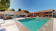 This outdoor amenity space centers around a generously sized, crystal-clear swimming pool with a connected spa, both finished with clean concrete surrounds and equipped with stainless steel entry rails. The pool deck offers ample room for lounging, furnished with teal-framed chaise lounge chairs arranged to take full advantage of the open sky and abundant natural sunlight. Teal umbrella-shaded patio tables and chairs provide comfortable spots for gathering and relaxation throughout the deck area.

A single-story clubhouse building with a pitched, brown shingled roof and large glass doors lines one side of the pool area, offering convenient interior access. Brick detailing and warm earth-toned exterior finishes carry throughout the surrounding multi-story residential buildings, which frame the space and create a cohesive, enclosed community feel. Decorative masonry pillar fencing with iron railings defines the perimeter of the pool area, maintaining both security and an open sightline to the surrounding landscaping.

Mature trees, including tall pines, are positioned throughout the grounds, providing natural shade and greenery against the backdrop of a clear blue sky. The overall atmosphere is bright, well-maintained, and inviting, designed to support outdoor recreation and social activity in a community setting.